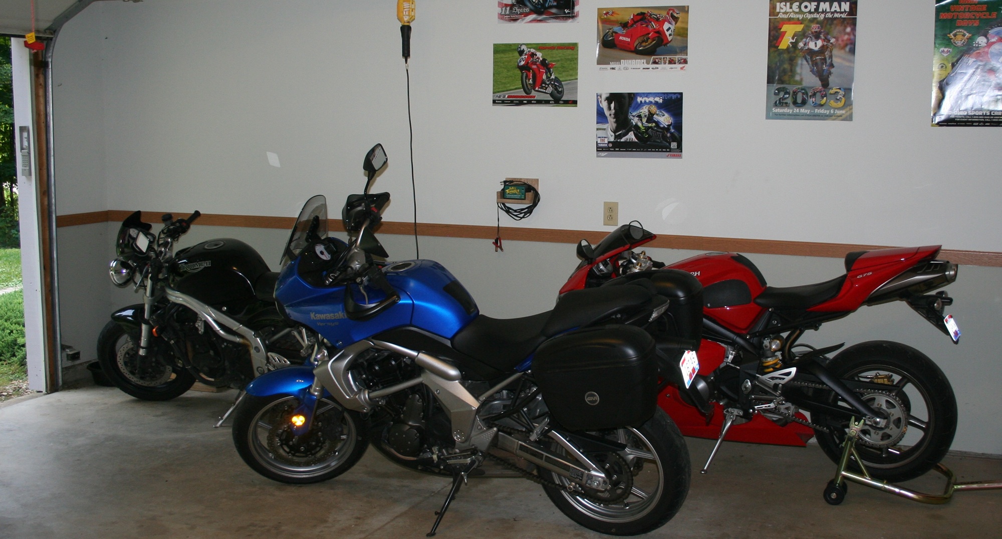 red Triumph Daytona sport bike, black Triumph Speed Triple, and blue Kawasaki Versys with saddlebags parked in a garage underneath racing posters