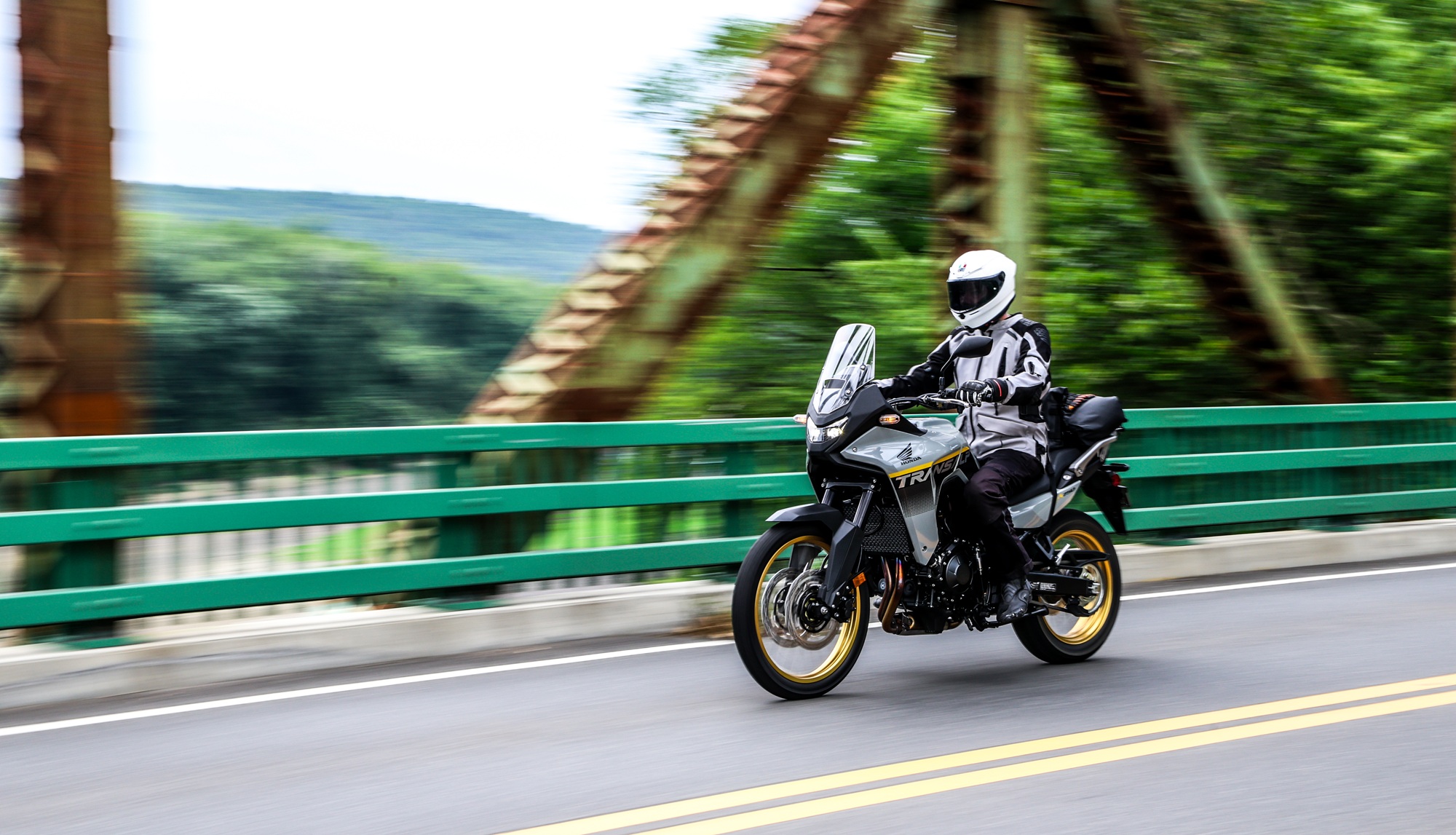 rider on the Transalp crossing an old rusty bridge with trees and a river in the background