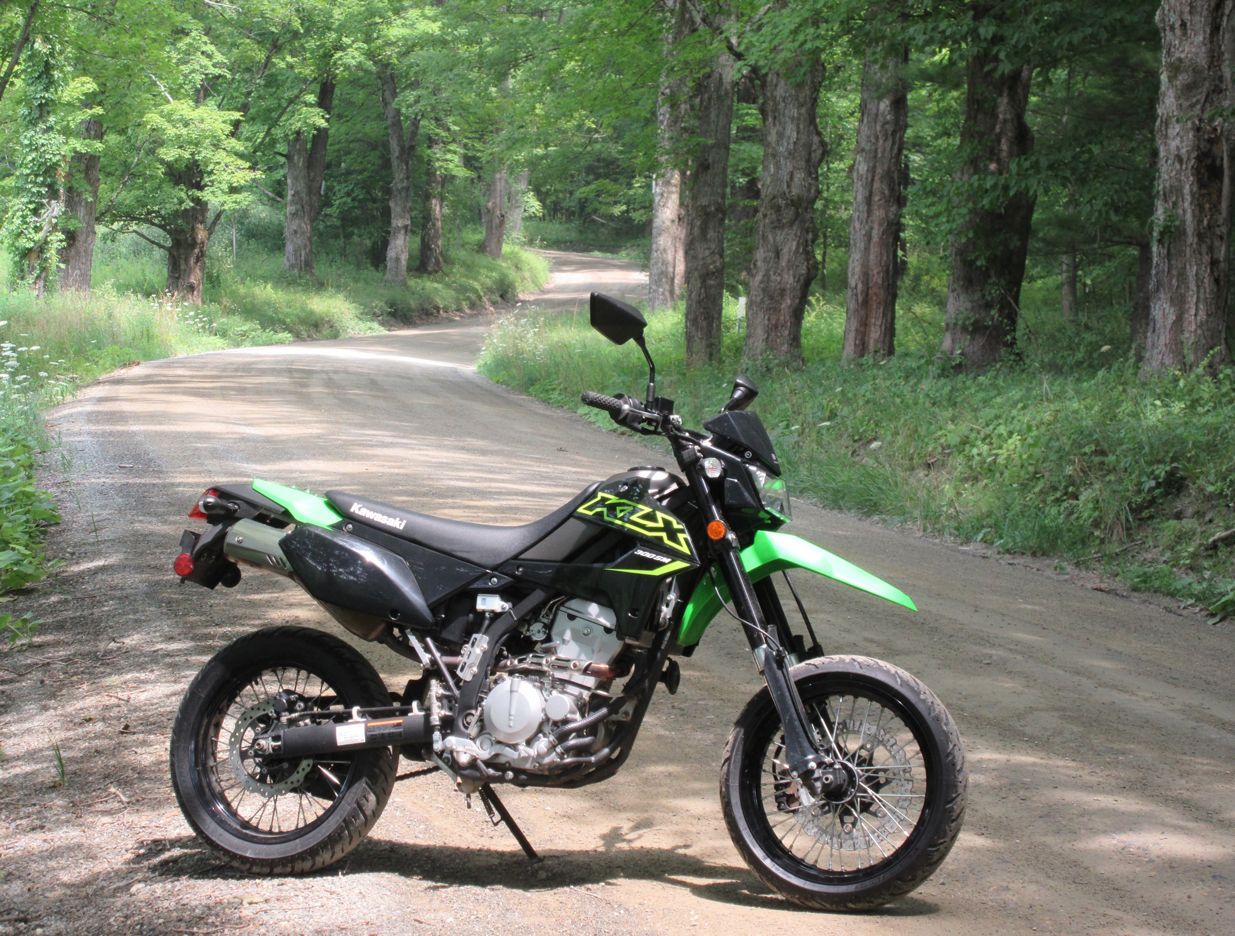 green and black supermoto KLX300SM stopped along a dirt road through a forest