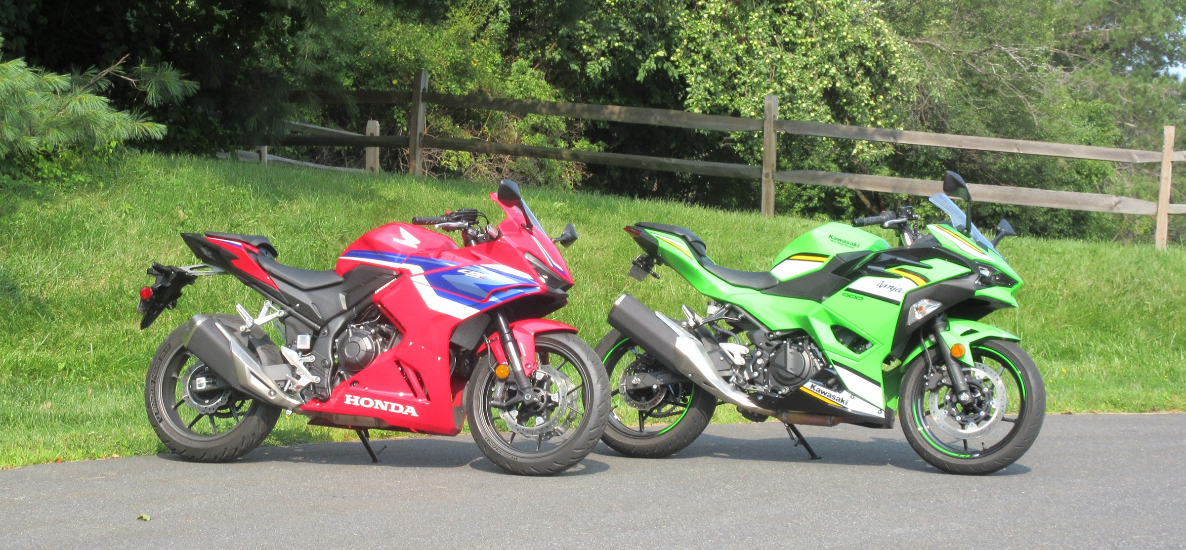 red CBR500R and green Ninja 500 parked side by side in front of trees and a split-rail fence