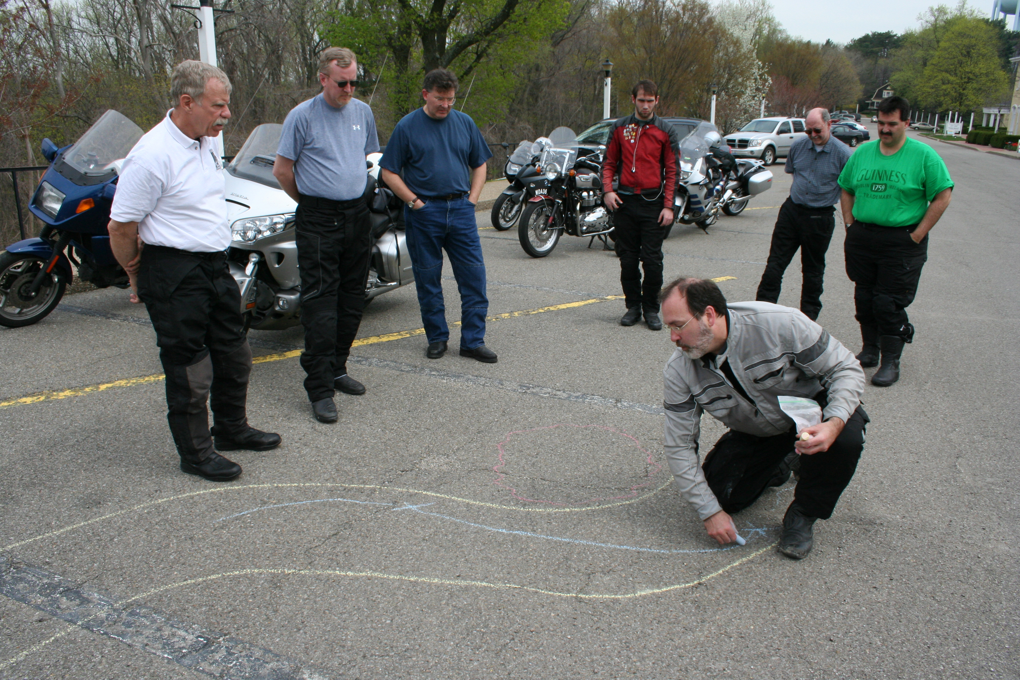 instructor drawing lines through a curve in a chalk drawing on pavement with students watching, motorcycles parked in the background