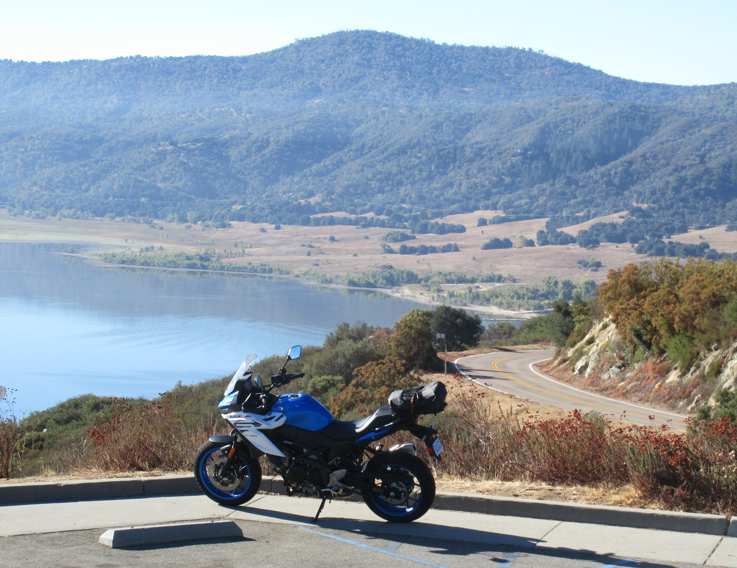 the blue and white version parked at an overlook above a lake with a curving road in the background and a dry bag strapped to the rack on the motorcycle