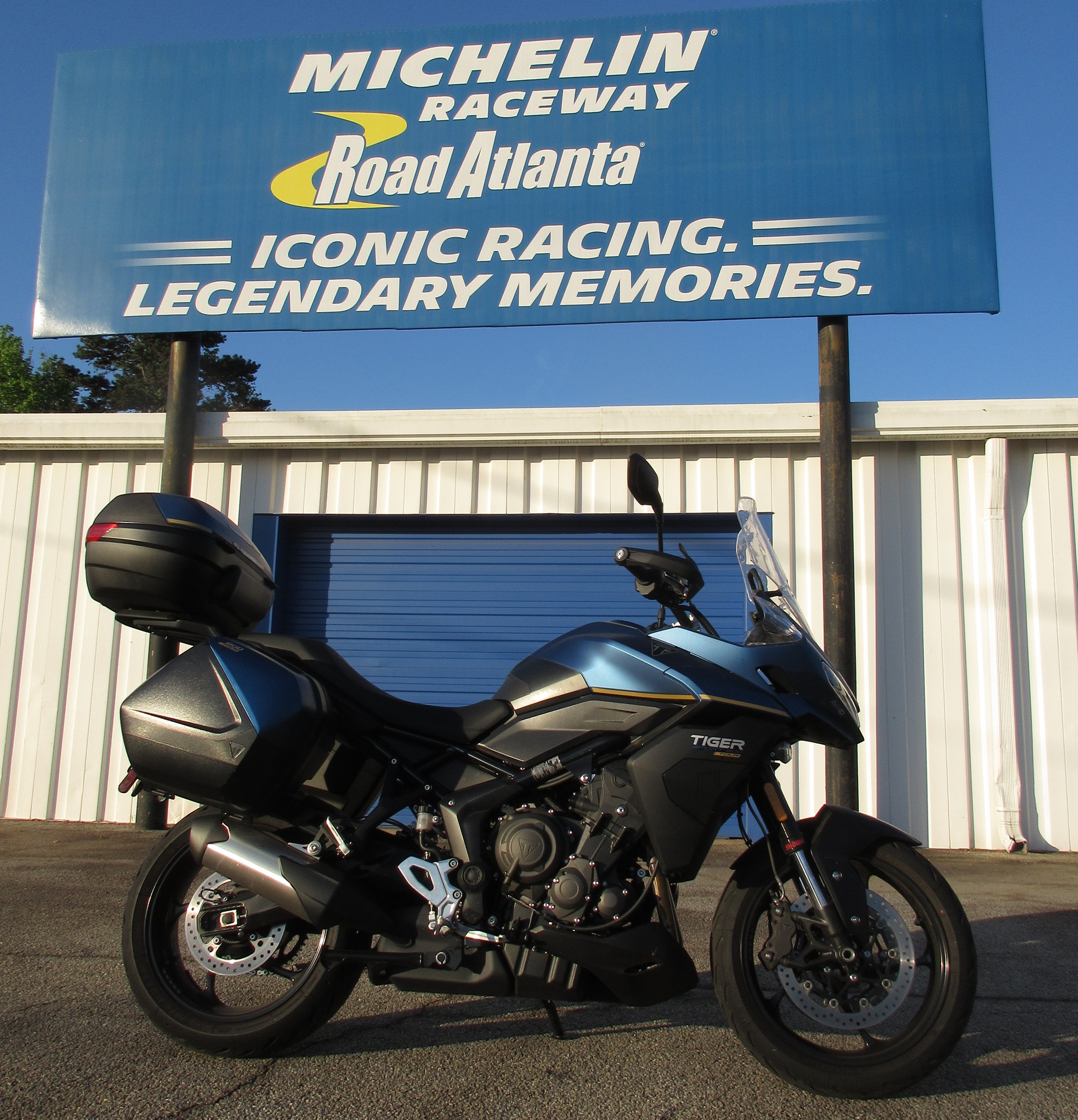 blue Triumph Tiger Sport 800 Tour motorcycle parked under a blue sign for Michelin Raceway Road Atlanta in front of garages