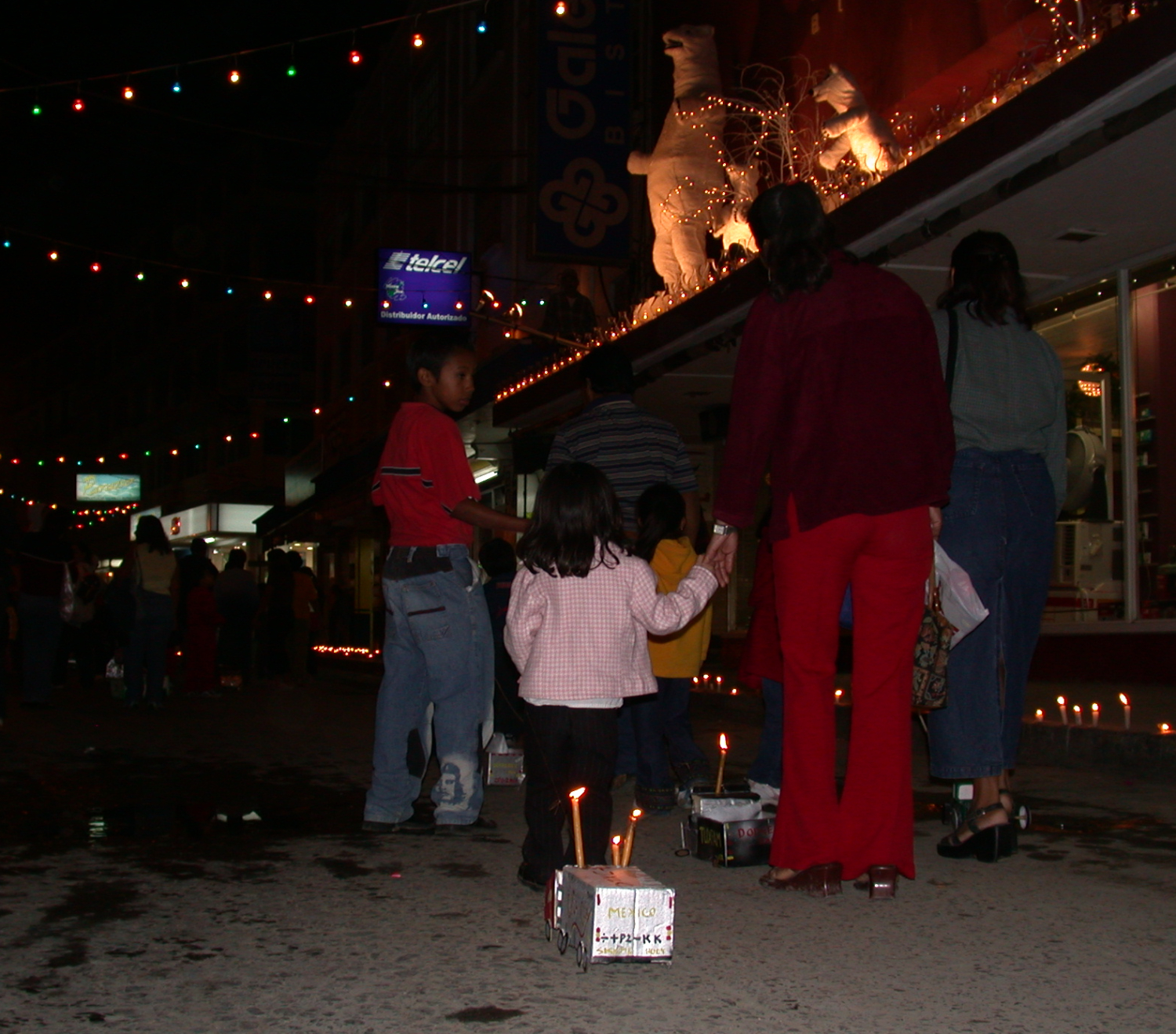 family walking hand in hand on a dark street with colored lights overhead with a girl pulling a small decorated car on a string