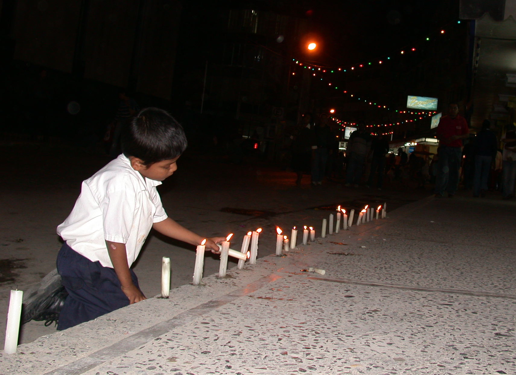 a child kneeling in the dark by a row of lit candles along a street, lighting one candle with another